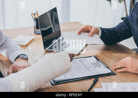 Vista ritagliata di imprenditore seduta a tavola e dando il foglio di carta al lavoratore con braccio rotto in ufficio, concetto di compensazione Foto Stock