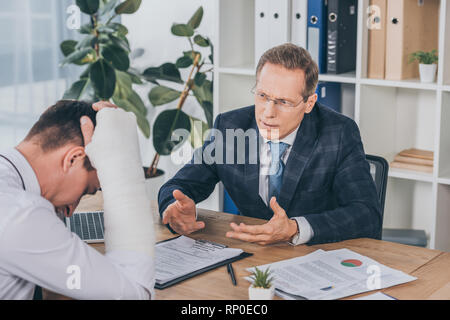 Lavoratore con braccio rotto seduta a tavola e la lettura dei documenti di fronte ai proprietari in giacca blu in ufficio, concetto di compensazione Foto Stock