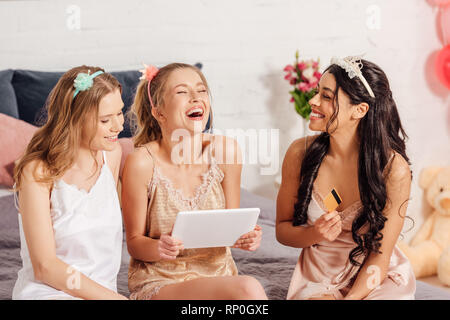 Bella sorridente ragazze multiculturale in calzature con tavoletta digitale e carta di credito facendo shopping online durante il pigiama party Foto Stock