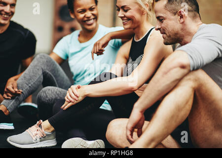 Gruppo di diversi amici in sportswear ridere mentre è seduto su un pavimento palestra dopo aver lavorato insieme Foto Stock