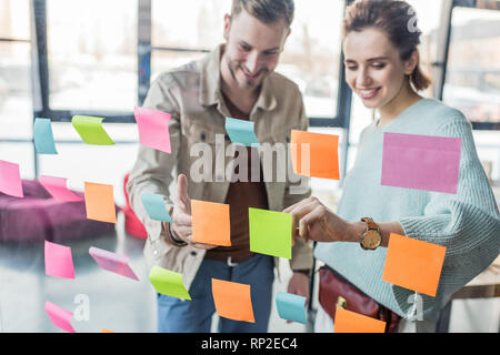 Sorridente imprenditore casual e imprenditrice mettendo colorato sticky notes sul vetro della finestra in ufficio Foto Stock