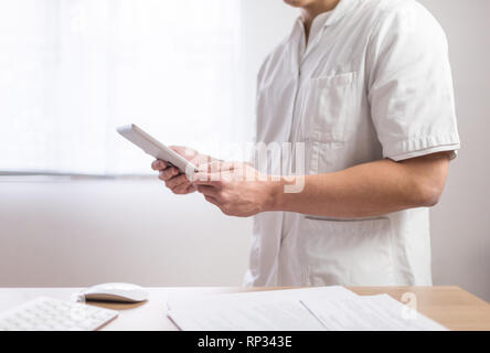 Medico nell'uniforme utilizzando computer tablet accanto alla sua scrivania in ufficio in ospedale Foto Stock