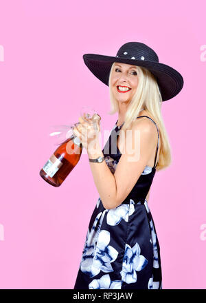 Felice, attraente donna bionda che porta una bottiglia di vino e un paio di bicchieri di vino Foto Stock