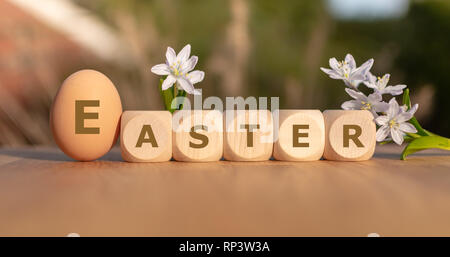 Un uovo e le piastrine formano la parola "Pasqua" su un tavolo in legno con fiori. Foto Stock