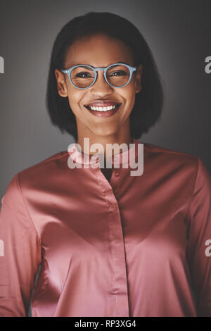 Sorridente giovane africana aziendale professionale con gli occhiali e sorridente con fiducia mentre in piedi contro uno sfondo grigio Foto Stock