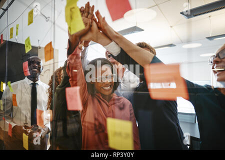 Eccitato gruppo di diversi imprenditori celebrando con alta cinque dopo una sessione di brainstorming con Sticky Notes in un ufficio Foto Stock
