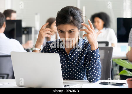 Sconvolto indian lavoratrice seduto alla scrivania in ufficio guardando lo schermo del PC si sente infelice e sconvolto. Millenaria donna indù lettura notific sgradevole Foto Stock