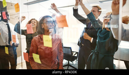 Il tifo gruppo di entusiasti imprenditori di celebrare un'idea vincente insieme durante il brainstorming con foglietti adesivi su una parete di vetro in un ufficio moderno Foto Stock