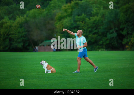 Bel giovane con Bulldog inglese cane all'aperto in città il parco forestale. Un uomo su un prato verde con il cane. Cynologist. Foto Stock