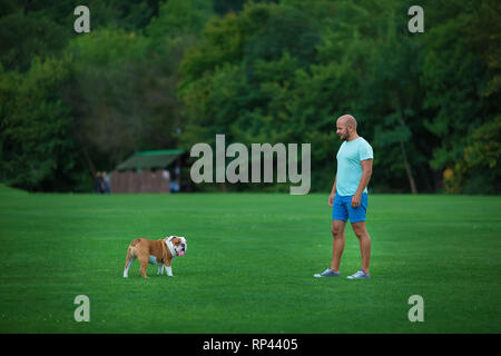 Bel giovane con Bulldog inglese cane all'aperto in città il parco forestale. Un uomo su un prato verde con il cane. Cynologist. Foto Stock