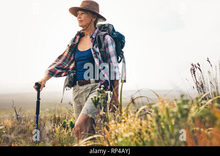 Senior donna su una escursione a piedi con l aiuto di un trekking pole. Woman trekking nella campagna a piedi attraverso le boccole. Foto Stock