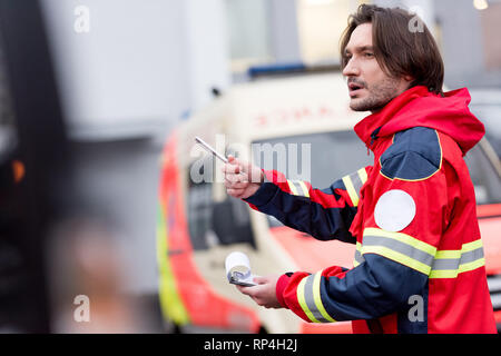 Brunette paramedico in rosso uniforme tenendo la penna e appunti su strada Foto Stock