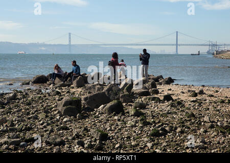 La gente sulla riva del fiume Tago sulla banchina delle colonne (Cais das Colunas) vicino alla Praça do Comércio a Lisbona, Portogallo. Il XXV Aprile Bridge (Ponte 25 de Abril) è visto in background. Foto Stock