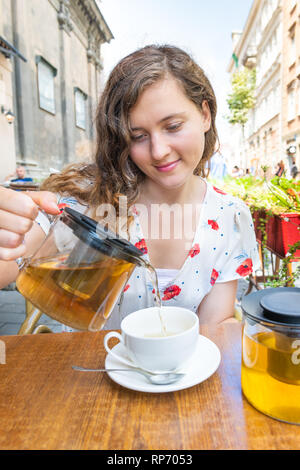 Ragazza giovane donna seduta nella Comunità outdoor cafe ristorante versando il tè verde nella tazza in estate a Lviv o Lvov, Ucraina città Foto Stock