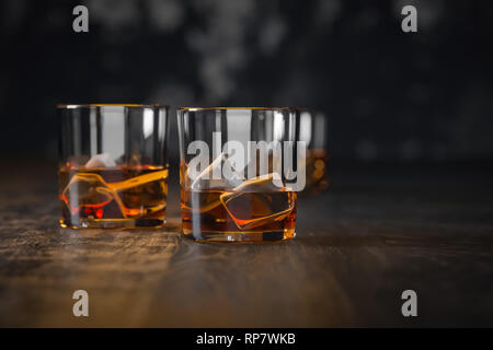 Whiskey tre bicchieri con ghiaccio, stand al buio su un vecchio tavolo in legno e uno sfondo nero Foto Stock