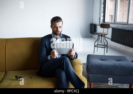 Lavorando...di successo e di moda uomo d affari è utilizzando una compressa mentre è seduto sul divano in un ufficio moderno. Aspetto di business Foto Stock
