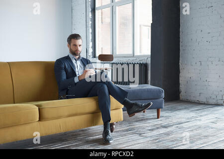 Look aziendale: successo e bello business man è seduto sul divano per ufficio e guardando a parte sul serio. Aspetto di moda. Concetto di successo Foto Stock