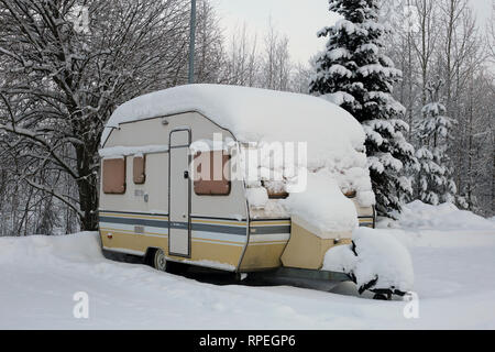 Caravan rimorchio in un parcheggio accanto a una foresta coperta di neve durante il periodo invernale in Europa. Il rimorchio è giallo e grigio colorato. Foto Stock