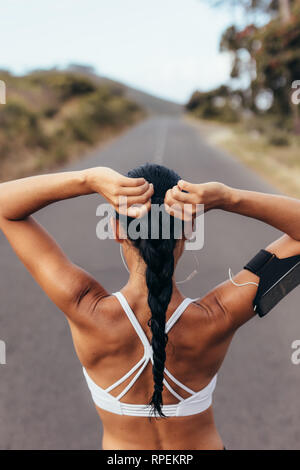 Vista posteriore del colpo di montare atleta femminile capelli di legatura prima del suo allenamento. Giovane donna ottenere pronto per la formazione all'aperto su una strada vuota. Foto Stock