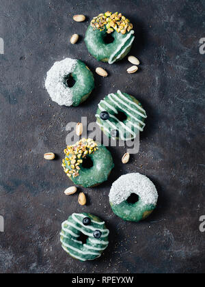 Vegan ciambelle con semi di Chia e rabboccato con sani spirulina smalto con pistacchio, noce di cocco essicata e mirtillo. Blu Verde spirulina ciambelle, su sfondo nero, vista dall'alto piatto o laici. In verticale Foto Stock