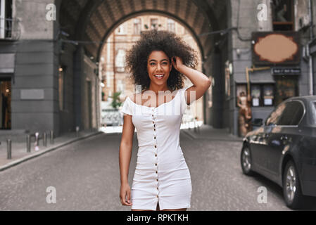 Bellezza senza sforzo. Attraente giovani Afro American donna in corto abito bianco a giocare con i capelli e sorridenti mentre in piedi all'aperto Foto Stock