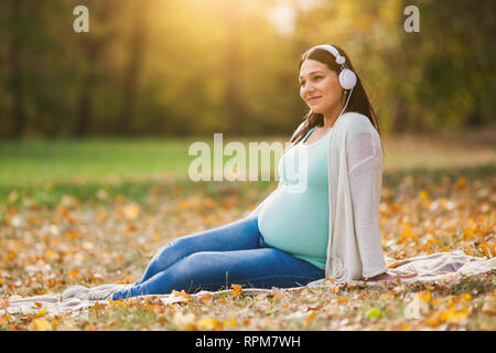 Donna incinta relax nel parco. Ella è in ascolto di musica. Foto Stock