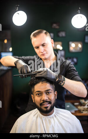 Parrucchiere taglia i capelli, con forbici e pettine nero, sulla corona del bello cliente soddisfatto su BARBER SHOP Foto Stock