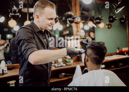 Styling professionale. Close up vista laterale del giovane uomo barbuto getting haircut dal parrucchiere con rasoio elettrico al barbiere Foto Stock