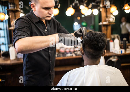 Styling professionale. Close up vista laterale del giovane uomo barbuto getting haircut dal parrucchiere con rasoio elettrico al barbiere Foto Stock