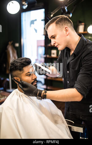 Styling professionale. Close up vista laterale del giovane uomo barbuto getting haircut dal parrucchiere con rasoio elettrico al barbiere Foto Stock