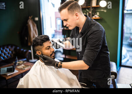 Styling professionale. Close up vista laterale del giovane uomo barbuto getting haircut dal parrucchiere con rasoio elettrico al barbiere Foto Stock