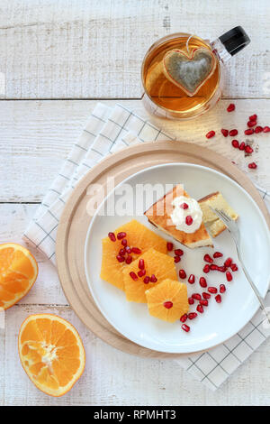 La colazione piatto composizione laici con un pezzo di torta, alcune fette di arancia e una tazza di tè con una forma di cuore un sacchetto da tè in esso Foto Stock