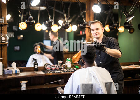 Barbiere con asciugacapelli e di asciugatura per lo styling dei capelli del cliente. Barbiere con asciugacapelli lavora sull'acconciatura per uomo barbuto, barbiere sfondo. Hipster bear Foto Stock