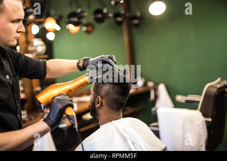 Barbiere con asciugacapelli e di asciugatura per lo styling dei capelli del cliente. Barbiere con asciugacapelli lavora sull'acconciatura per uomo barbuto, barbiere sfondo. Hipster bear Foto Stock