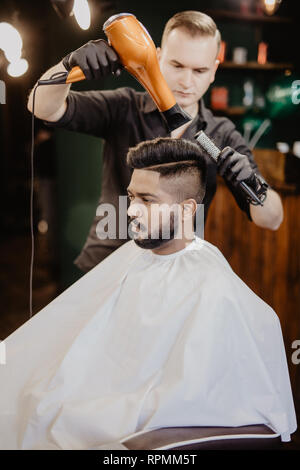 Barbiere con asciugacapelli e di asciugatura per lo styling dei capelli del cliente. Barbiere con asciugacapelli lavora sull'acconciatura per uomo barbuto, barbiere sfondo. Hipster bear Foto Stock