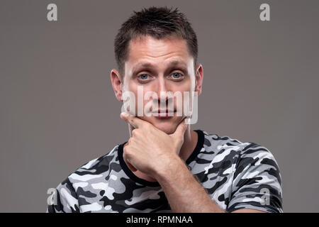 Ritratto di incerta espressiva uomo guardando la fotocamera con le mani sul mento su sfondo grigio. Foto Stock