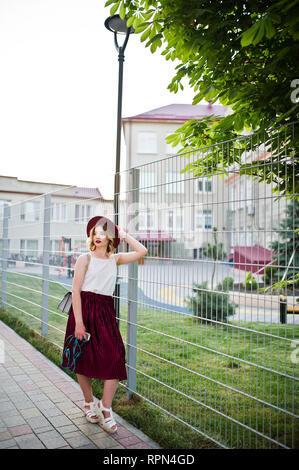 Elegante e bella bionda ragazza modello in elegante velluto rosso mantello in velluto, camicia bianca e cappello, poste con telefono e auricolari. Foto Stock