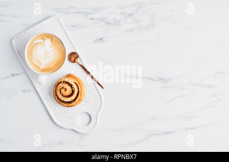 Pane appena sfornato cannella bun con spezie e riempimento di cacao e caffè o cappuccino con latte art su bianco che serve la piastra al di sopra di marmo bianco dello sfondo. Foto Stock