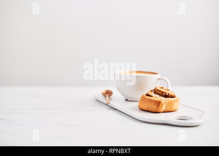 Pane appena sfornato cinnamon roll con spezie e riempimento di cacao e caffè o cappuccino bianco sul piatto di portata su marmo bianco tabella su sfondo bianco. Foto Stock