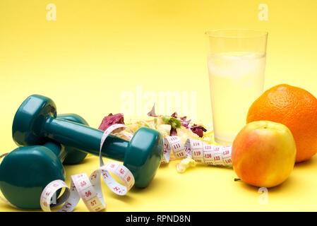 Pesi per lo sport. Insalata di fitness e di misura di nastro su una tabella di colore giallo. Arance e mele e acqua chiara con ghiaccio per il concetto di una sana lifestyl Foto Stock