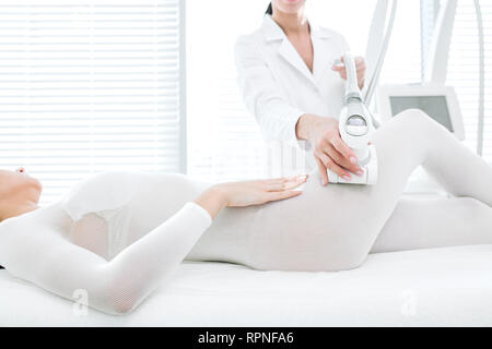 Irriconoscibile estetista femminile in medicina bianco vestaglia assiste paziente di sesso femminile in cosmetologico wellness clinic, esecuzione di massaggio GPL sui fianchi in w Foto Stock