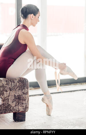 Giovane ballerino slim mettendo sul pointe scarpe mentre seduto sul banco. vista laterale a piena lunghezza foto. Foto Stock
