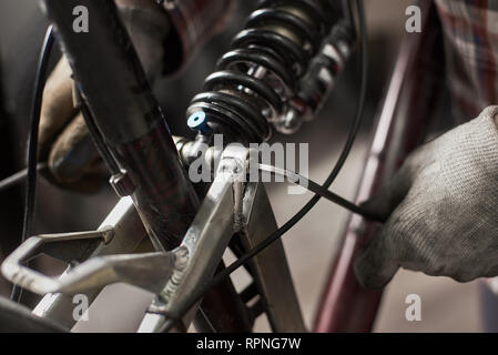 Vista ritagliata di maschio meccanico che lavora nel negozio di riparazione di biciclette, riparazione bici utilizzando lo strumento speciale, indossare guanti di protezione Foto Stock