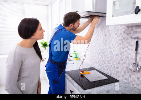 Repairman maschio estrattore di controllo filtro con multimetro digitale mentre la donna in piedi in cucina Foto Stock