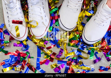 Due piedi in scarpe bianche tra festa multicolore confetti sul pavimento grigio Foto Stock