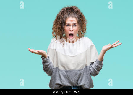 Bella bruna capelli ricci giovane ragazza con gli occhiali su sfondo isolato celebrando mad e pazzo per il successo con le braccia sollevate e chiuso ey Foto Stock