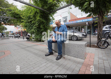 Un balding di mezza età, sub-continente l uomo nel suo late 40's siede concentrare durante la lettura dal suo telefono in John Street Cabramatta a Sydney Foto Stock