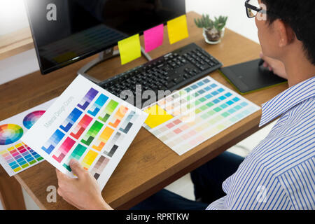 Editor di designer al lavoro disegno tratteggia un nuovo progetto sulla tavoletta grafica e la tavolozza dei colori seduto alla scrivania con computer in studio o in ufficio. Foto Stock