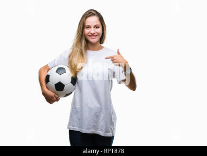 Giovane bella donna bionda tenendo palla calcio su sfondo isolato con sorpresa faccia puntare il dito a se stesso Foto Stock