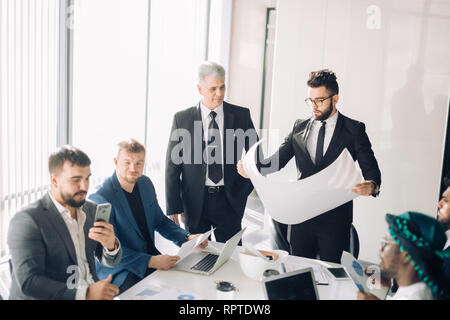 Handome maschio caucasico manager rende la lavagna a fogli mobili la presentazione di diversi colleghi alla riunione Foto Stock
